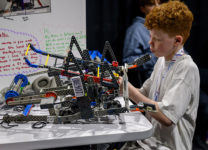 A boy examines a VEX Robotics robot on a table, with a handwritten team introduction poster visible in the background.
