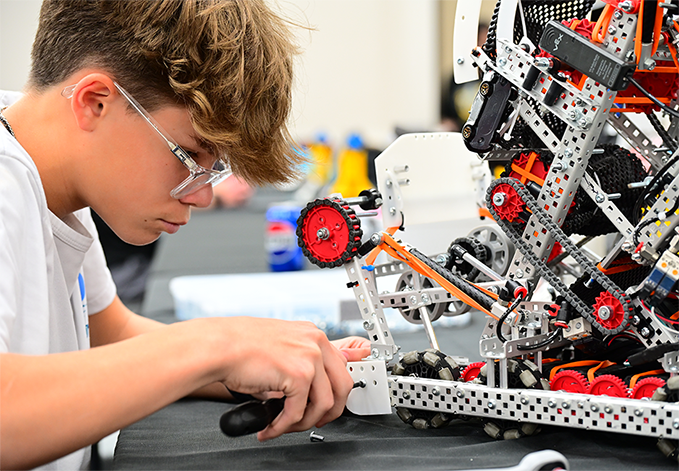 A teenage boy wearing safety glasses carefully adjusts components on a tall, complex VEX robotics build at a workbench.