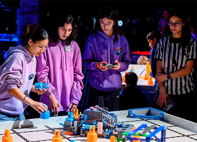 Three students in purple hoodies operate a VEX robotics robot on a competition field, with a referee in a striped shirt observing nearby.
